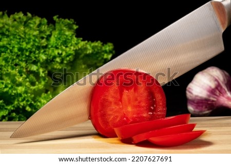 A metal knife slicing a red ripe tomato on a wooden board. Close up of a sharp knife blade cutting tomato on slices, green salad and garlic on a black isolated background. Royalty-Free Stock Photo #2207627691