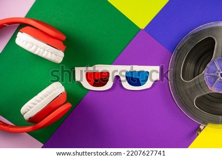 Old white paper 3d glasses with blue and red lenses and red headphones on vintage color background. Retro stereoscopic 3D glasses and filmstrip spool on green, purple, yellow and blue background.