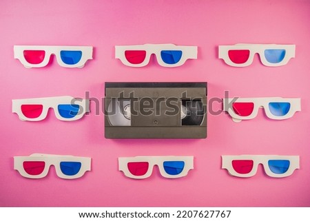Old white paper 3d glasses with blue red lenses and black videotape on pink background. Retro video cassette with magnetic tape and stereoscopic 3D cardboard glasses. Vintage background.
