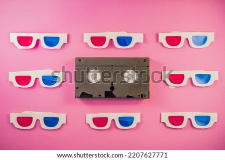Old white paper 3d glasses with blue red lenses and black videotape on pink background. Retro video cassette with magnetic tape and stereoscopic 3D cardboard glasses. Vintage background.