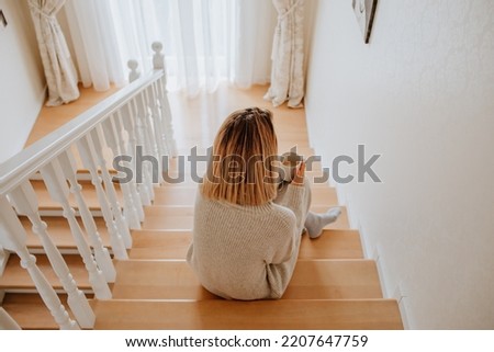 Pretty young woman in long winter dress is sitting on the stairs in the house and feeling bad and cold. Social media and healthcare concept. View from the back Royalty-Free Stock Photo #2207647759
