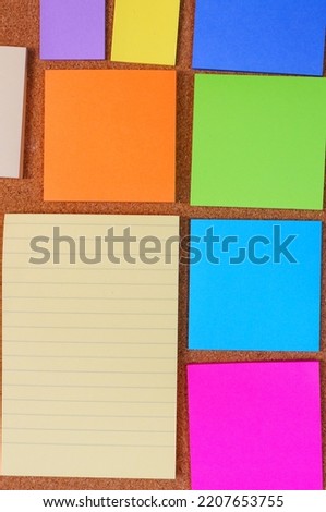 Yellow and colorful cards for the office, stickers for memory - unprinted, along with a piece of a large office notebook, on the background of a cork board. 