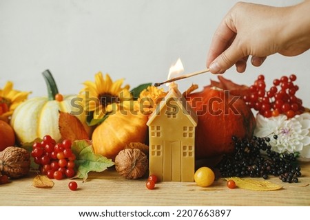 Hand lighting up candle on background of stylish pumpkins, autumn flowers, berries, leaves on wooden table in rustic room. Atmospheric autumn banner. Happy Thanksgiving! Seasons greeting card