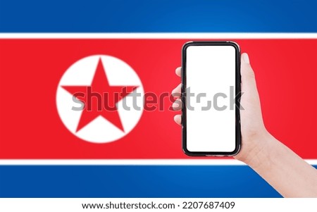 Close-up of male hand holding smartphone with blank on screen, on background of blurred flag of North Korea.