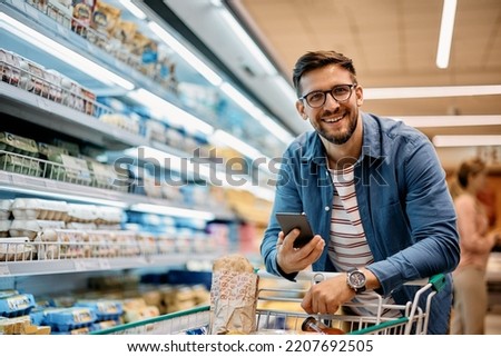 Happy man using smart phone while shopping in supermarket and looking at camera.