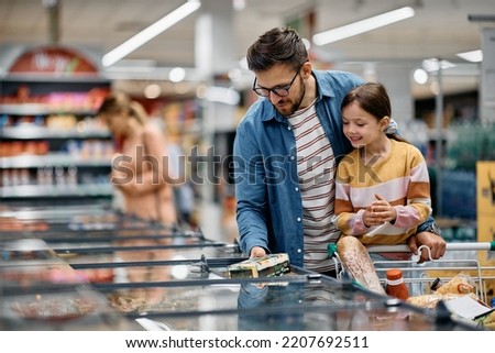 Happy little girl and her dad choosing food at refrigerated section while shopping in supermarket. Royalty-Free Stock Photo #2207692511