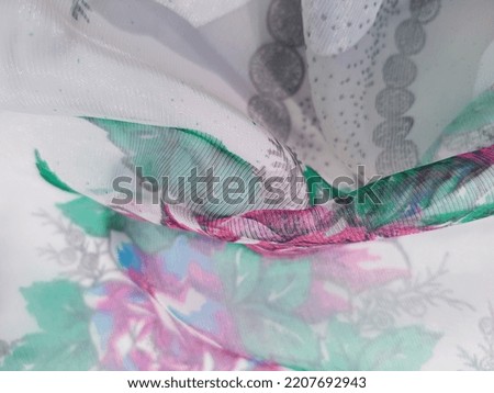 Wavy translucent slightly shiny light fabric (organza) with a floral print of silhouettes of pink and blue flowers in green leaves (macro, texture).