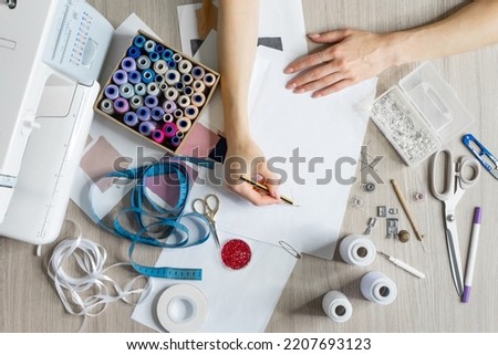 Women's hands draw sketches of dresses with pencil. Top view of sewing items and seamstress tools. Tailoring scissors, measuring tape, sewing machine, threads, beads, buttons, fabric, awl. 