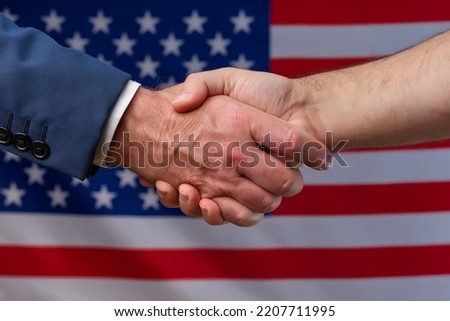 Two men shaking hands with the United States flag in the background. Cooperation and political protection of citizens in America. Alliance with the USA Royalty-Free Stock Photo #2207711995
