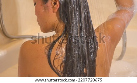 CLOSE UP: Back view shot of beautiful woman rinsing her hair with shower handle. Pretty young lady showering her dark long hair while sitting in bathtub. Time for personal hygiene in home bathroom. Royalty-Free Stock Photo #2207720675