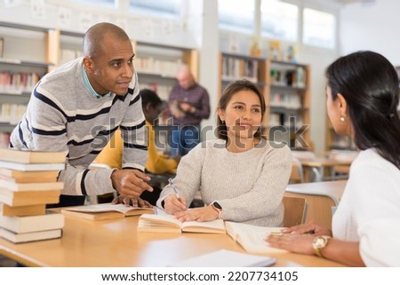 Group of positive adult hispanic students communicating in library, talking, reading books and making notes
