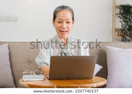 Happy asian mature woman remote working on laptop, sitting on couch.