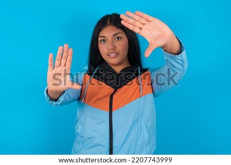 Portrait of smiling Young latin woman wearing sport clothes over blue background looking at camera and gesturing finger frame. Creativity and photography concept.
