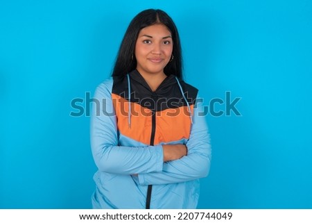 Waist up shot of  self confident Young latin woman wearing sport clothes over blue background has broad smile, crosses arms, happy to meet with colleagues.