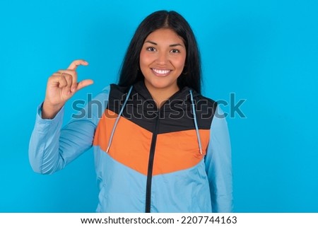 Young latin woman wearing sport clothes over blue background  smiling and gesturing with hand small size, measure symbol.