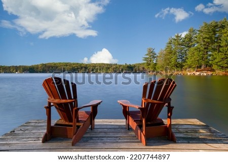 Two Adirondack chairs on a wooden dock overlooking a calm lake.  Cottages nestled between green trees are visible across the water. Royalty-Free Stock Photo #2207744897