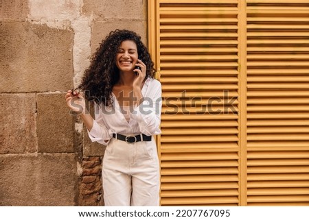 Happy young african woman laughing leaned on wall talking on her phone during spring day. Brunette hair girl wears casual clothes. Concept modern technologies. 