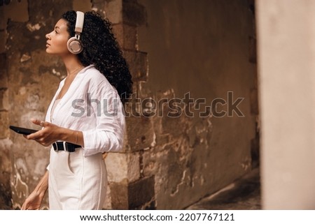 Beautiful young african woman with headphones using her smartphone, stands on old europe block. Girl with curly brunette hair wears shirt and white jeans. Concept telephone technology.