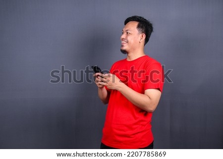 Portrait of a young cheerful asian man wearing red t-shirt standing isolated over gray background, using mobile phone