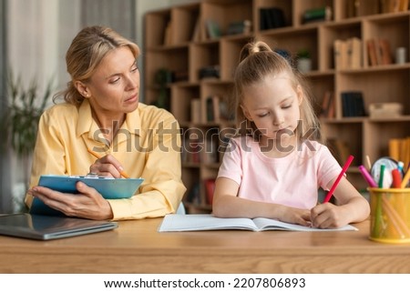 Art development concept. Talented little girl drawing picture with colorful pencils, woman psychologist watching kid and taking notes at clipboard, sittign at desk at office