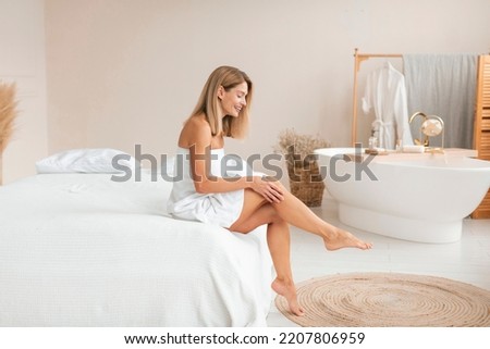 Beautiful middle aged woman in towel touching her leg with soft skin after depilation, sitting on bed at home, copy space. Mature lady after hair removal procedure, full length Royalty-Free Stock Photo #2207806959