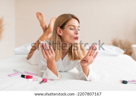 Gorgeous middle aged woman doing manicure, applying nail polish and blowing on nails while lying on bed at home. Mature lady making self-care procedure indoors. Domestic spa salon Royalty-Free Stock Photo #2207807167