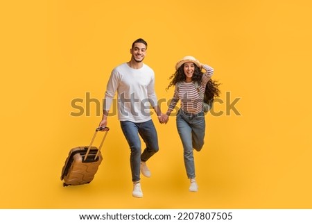 Portrait Of Cheerful Young Arab Couple Jumping With Suitcase On Yellow Background, Happy Middle Eastern Man And Woman In Straw Hat Laughing And Smiling At Camera, Enjoying Travel And Vacation