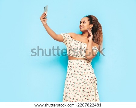 Beautiful woman with curls hairstyle. Smiling model dressed in summer dress. carefree female posing near blue wall in studio. Tanned and cheerful. Taking selfie