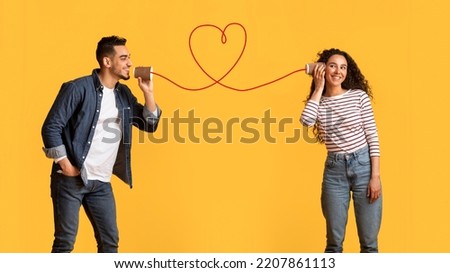 Message With Love. Happy Young Arab Man Telling Romantic Words To His Girlfriend Through Tin Phone With Drawn Red Heart As String, Loving Middle Eastern Couple Posing Over Yellow Background Royalty-Free Stock Photo #2207861113