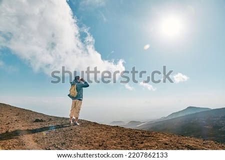 Rear view of man looking through binoculars at panoramic view of colorful summits of active volcano Etna, Tallest volcano in Continental Europe, Sicily, Italy. Royalty-Free Stock Photo #2207862133