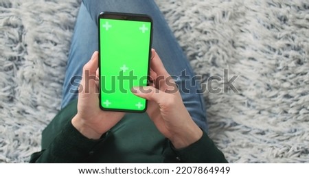 Female holds smartphone with green screen in hands. Top view, zoom in close up, living room, sofa. Using the app, swipe up. Unrecognizable woman.