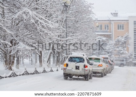Cars drive along a snow-covered city street. Heavy snowfall and snowstorm in the city. A lot of snow on the roadway, cars and trees. Cold snowy winter weather. Magadan, Siberia, Russia. Royalty-Free Stock Photo #2207871609