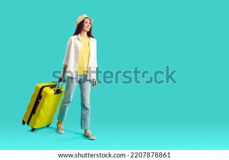 Joyful happy young female tourist or vacationer with suitcase goes on summer trip. Smiling girl in summer casual clothes walks with suitcase near copy space on light blue background. Full length.