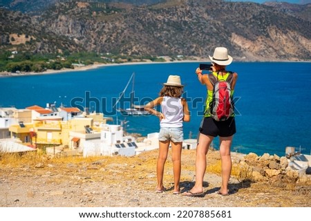 Tourists photographing a sailboat boat in Paleochora, Crete, Greece. Mother and daughter standing over Paleochora bay, taking pictures of the bay and ship with a phone. Crete, Greece.