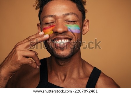 Cheerful African American gay smiling with closed eyes and painting rainbow on cheeks during LGBT pride event against brown background Royalty-Free Stock Photo #2207892481