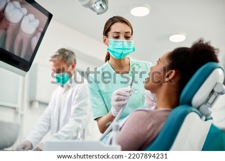 Nurse with face mask using suction tube while preparing African American woman for treatment at dental clinic. Royalty-Free Stock Photo #2207894231