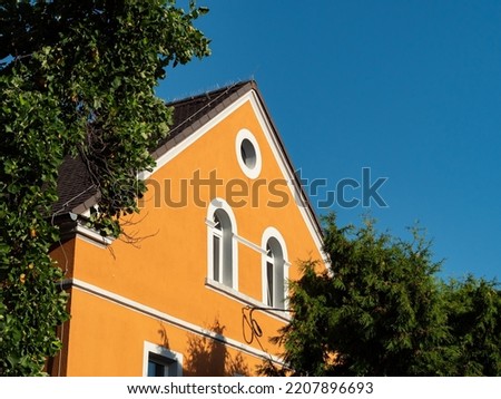An German house facade in between green tree crowns. Old building exterior with a gable roof in front of a blue sky. Ancient architecture in East Germany in a rural area. Renovated with orange color. Royalty-Free Stock Photo #2207896693