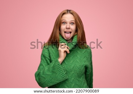 Cheerful young female with long ginger hair touching collar of green sweater and showing tongue while making funny faces against pink background