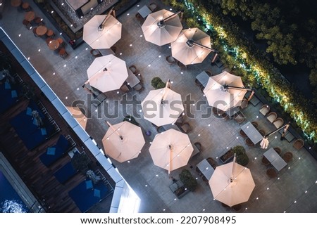A night top view of an esplanade of a resort cafe or a restaurant with sunshade umbrellas, tables and chairs, recliners next to the swimming pool, and an empty bar surrounded by illuminated trees Royalty-Free Stock Photo #2207908495