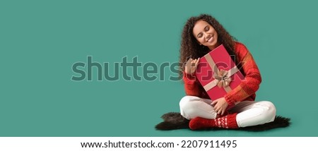 Young African-American woman in warm sweater holding Christmas gift on green background with space for text