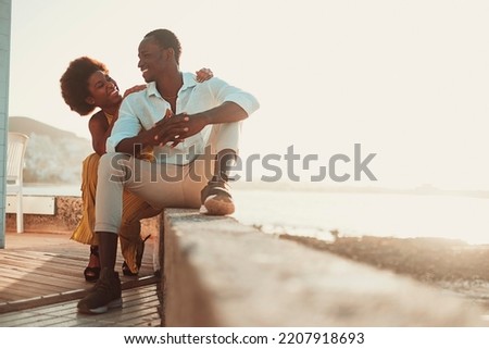 Black man and woman sitting and enjoying together outdoor leisure activity and dating. Happy young couple in love and friendship with sunny beach and sunset in background. Concept of summer people Royalty-Free Stock Photo #2207918693