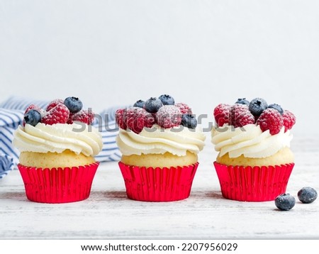 Vanilla cupcakes with cream cheese frosting and fresh berries in red cups. Beautiful colorful dessert for party or celebration, festive baking. White wooden background. Close up view. Copy space.