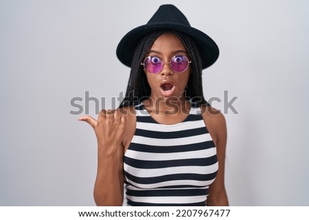 Young african american with braids wearing hat and sunglasses surprised pointing with hand finger to the side, open mouth amazed expression. 