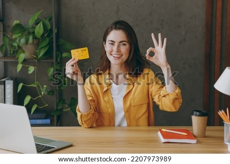 Young successful employee business woman 20s wear casual yellow shirt hold in hand mock up of credit bank card show ok gesture sit work at wooden office desk with pc laptop. Achievement career concept Royalty-Free Stock Photo #2207973989