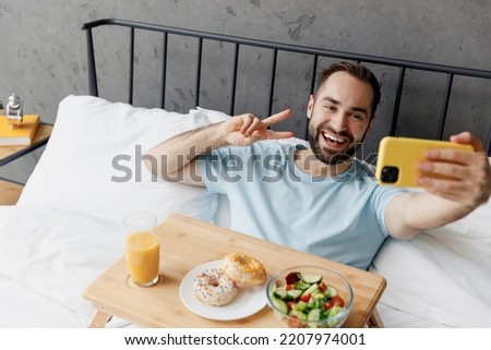 Young man in blue t-shirt lying in bed have breakfast doing selfie shot on mobile cell phone show thumb up rest relax spend time in bedroom lounge home in own room hotel. Good mood bedtime concept.