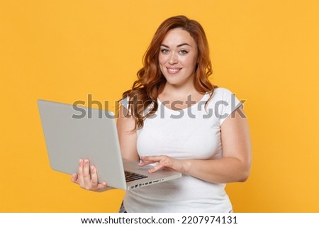 Smiling young redhead plus size body positive female woman girl 20s in white blank design casual t-shirt posing working on laptop pc computer isolated on yellow color wall background studio portrait