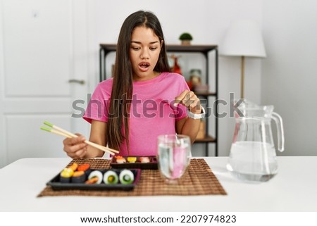 Young brunette woman eating sushi using chopsticks pointing down with fingers showing advertisement, surprised face and open mouth 