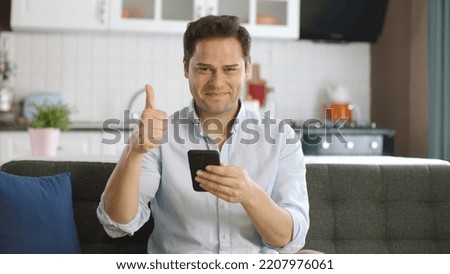Man using smartphone on sofa at home showing thumbs up sign as if he did a good job. Young man mobile technology user working on digital apps, searching information online, texting at home or office.