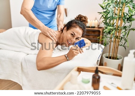 Middle age man and woman wearing therapist uniform having back massage session and holding credit card at beauty center