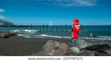 Woman in red swimming suit sitting on the rock in the black ocean sandy beach. Calm ocean water and pure blue sky at the background.